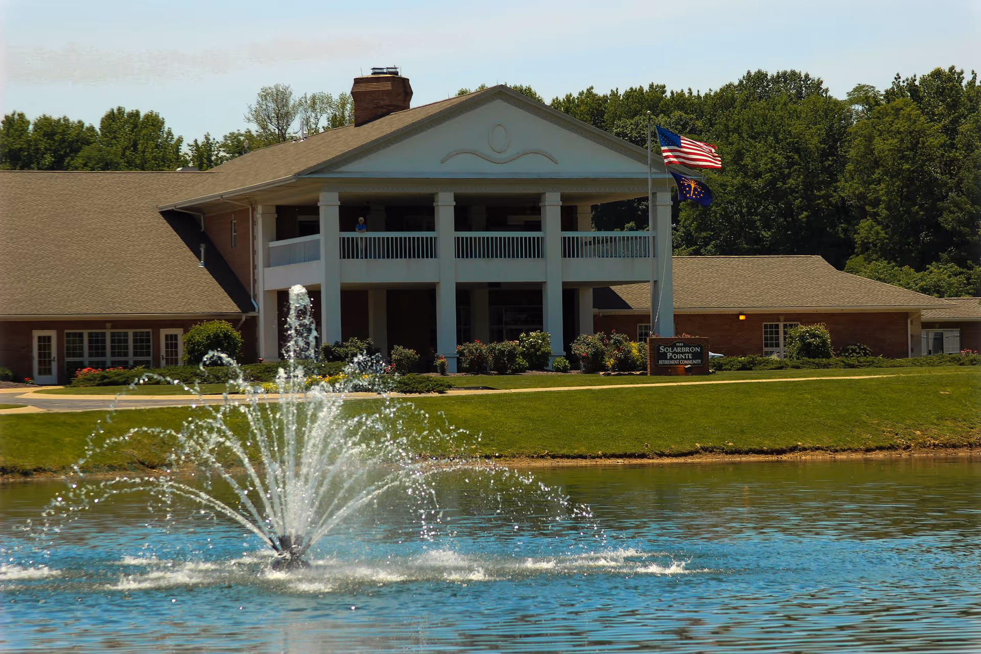 Two-story brick senior living building with white columns and a balcony, flags on a flagpole, and a fountain spraying in a pond in the foreground.