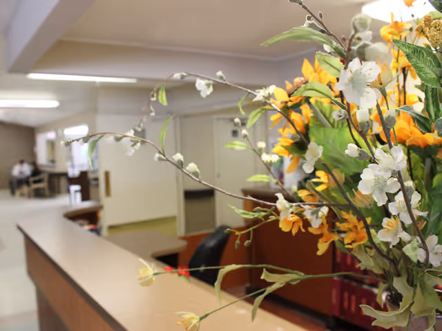 Interior view of a healthcare facility reception area with a wooden counter in the foreground and a bouquet of yellow and white flowers on the right side. In the background, there are doors, a hallway, and a few people sitting at tables.