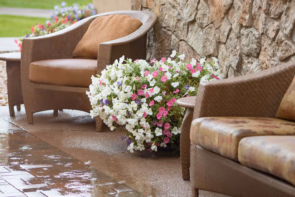 Outdoor seating area with brown cushioned wicker chairs and a wicker sofa next to a stone wall, with a large planter of white, pink, and purple flowers on a wet tiled floor.