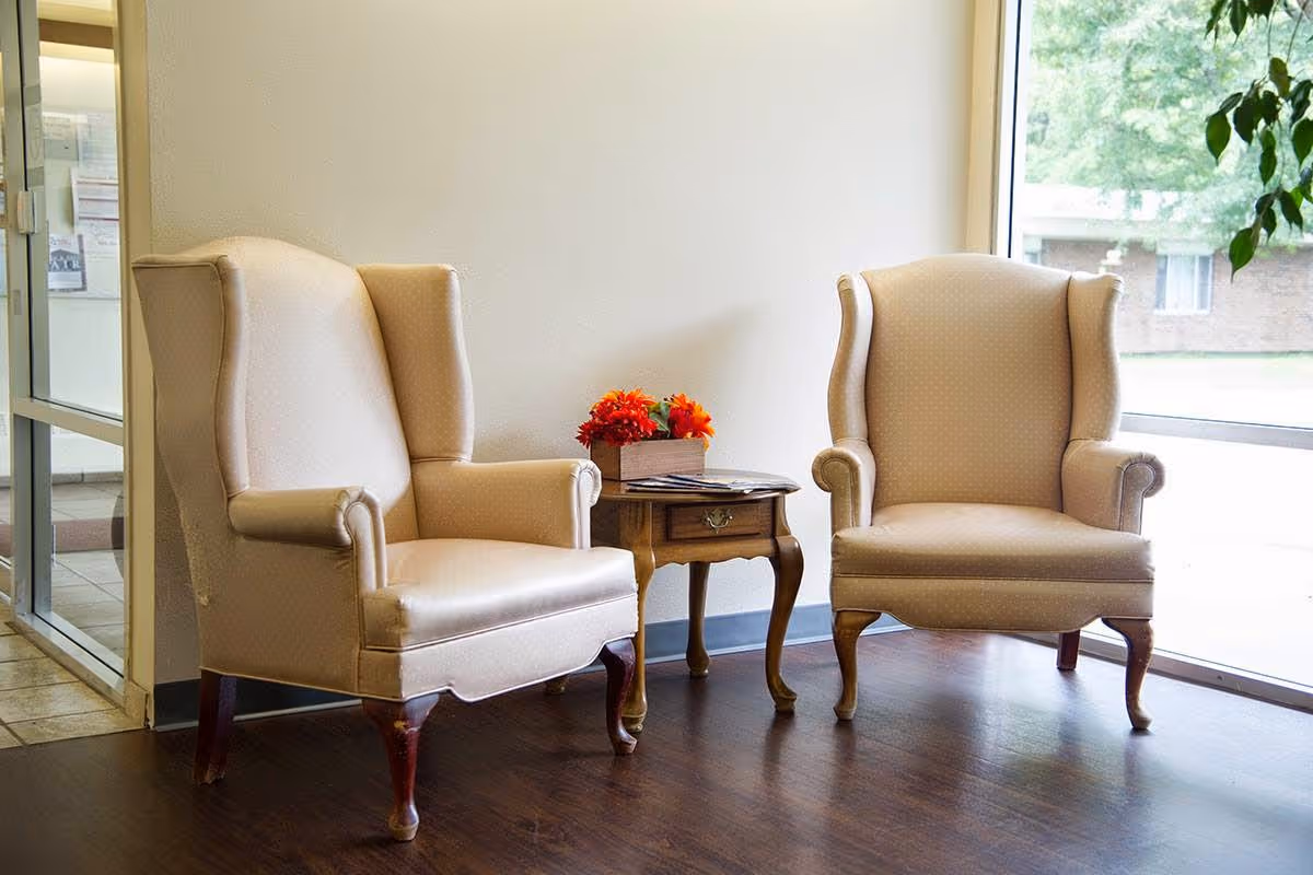 Two beige upholstered wingback chairs with wooden legs are positioned on either side of a small wooden side table with a drawer. On the table, there is a small arrangement of orange flowers and some magazines. The setting is indoors with a large window on the right side letting in natural light and a glass door on the left side.
