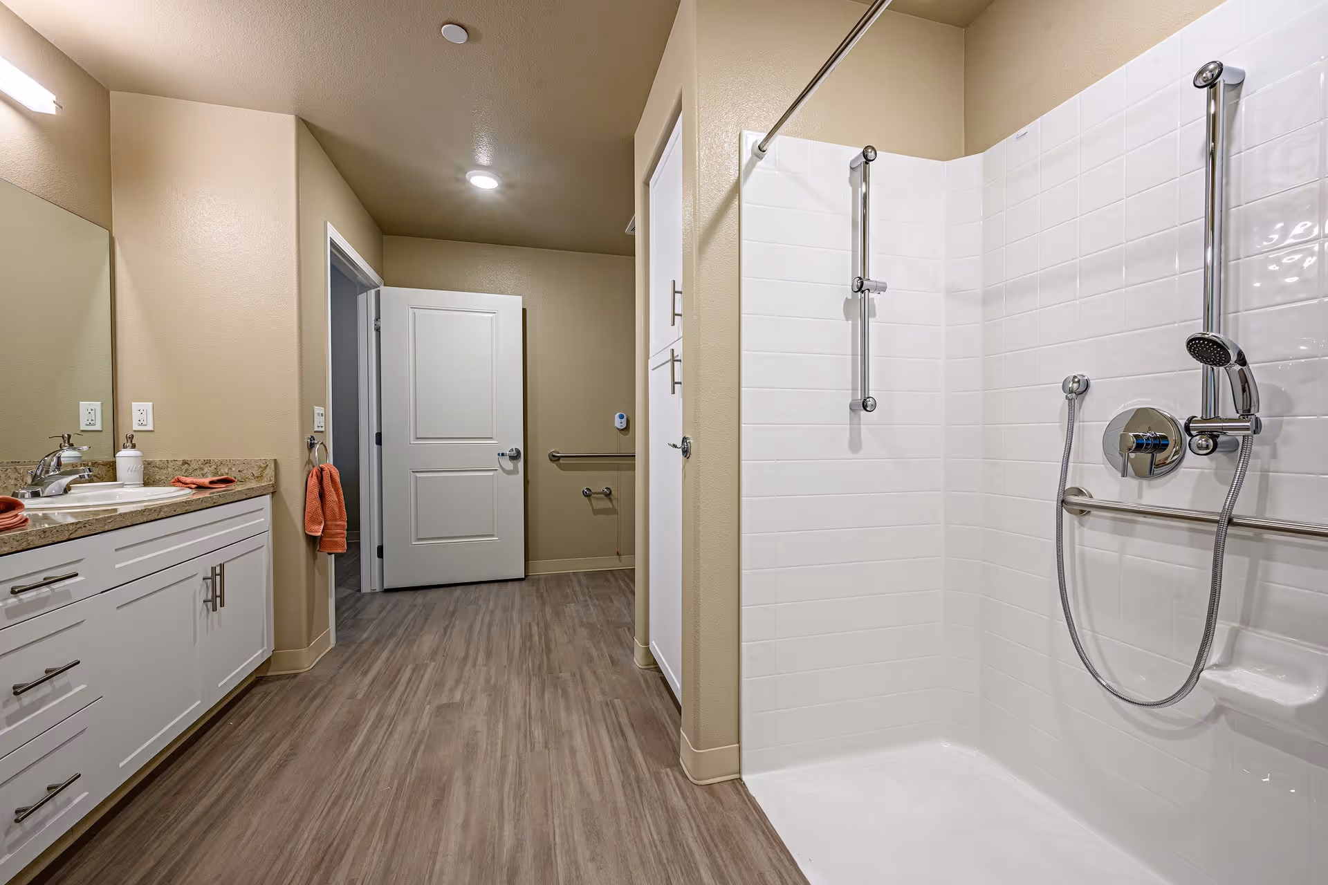 A modern bathroom with beige walls and wood-look flooring. On the left side, there is a white vanity with a granite countertop, a sink, a mirror, and orange towels. On the right side, there is a walk-in shower with white tiled walls and chrome fixtures, including a handheld showerhead and grab bars. A white door is open in the background, revealing a hallway with additional grab bars.