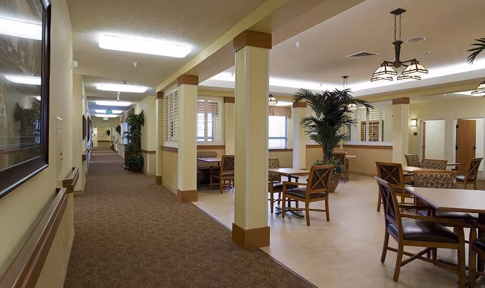 Interior view of a senior living facility hallway leading to a common area with tables and chairs. The space is well-lit with ceiling lights and decorated with potted plants and framed artwork on the walls. The common area has multiple seating arrangements with wooden chairs and tables, and windows with blinds allowing natural light.