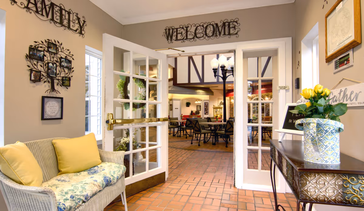 Interior view of a welcoming senior living facility entrance area with open double glass doors leading to a common seating area with tables and chairs. A wicker bench with yellow cushions is on the left, and a decorative table with a vase of yellow flowers is on the right. Wall decorations include a family photo tree and a welcome sign above the doors.