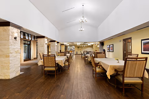Spacious dining room with multiple tables covered in beige tablecloths and wooden chairs with cushioned seats. The room features a high vaulted ceiling with chandeliers, stone pillars, wooden flooring, and framed artwork on the walls.