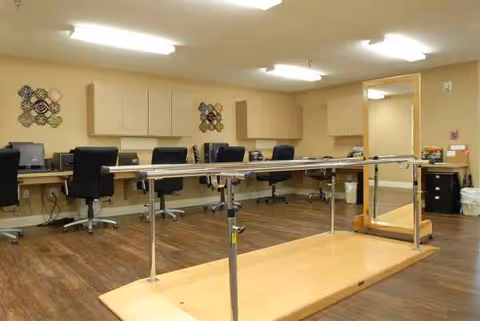 Therapy room with parallel bars, desks, and chairs along beige walls inside a senior living facility.