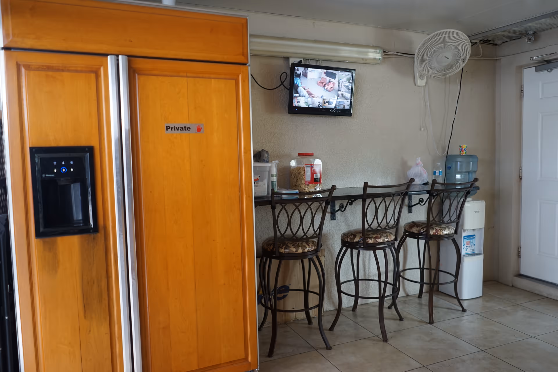 A small seating area with three high chairs lined up against a wall-mounted counter. Above the counter is a small TV screen showing multiple security camera feeds. To the left is a large wooden refrigerator with a 'Private' sign on one door. On the right side, there is a water cooler and a wall-mounted fan above it. The floor is tiled and there is a white door on the far right.