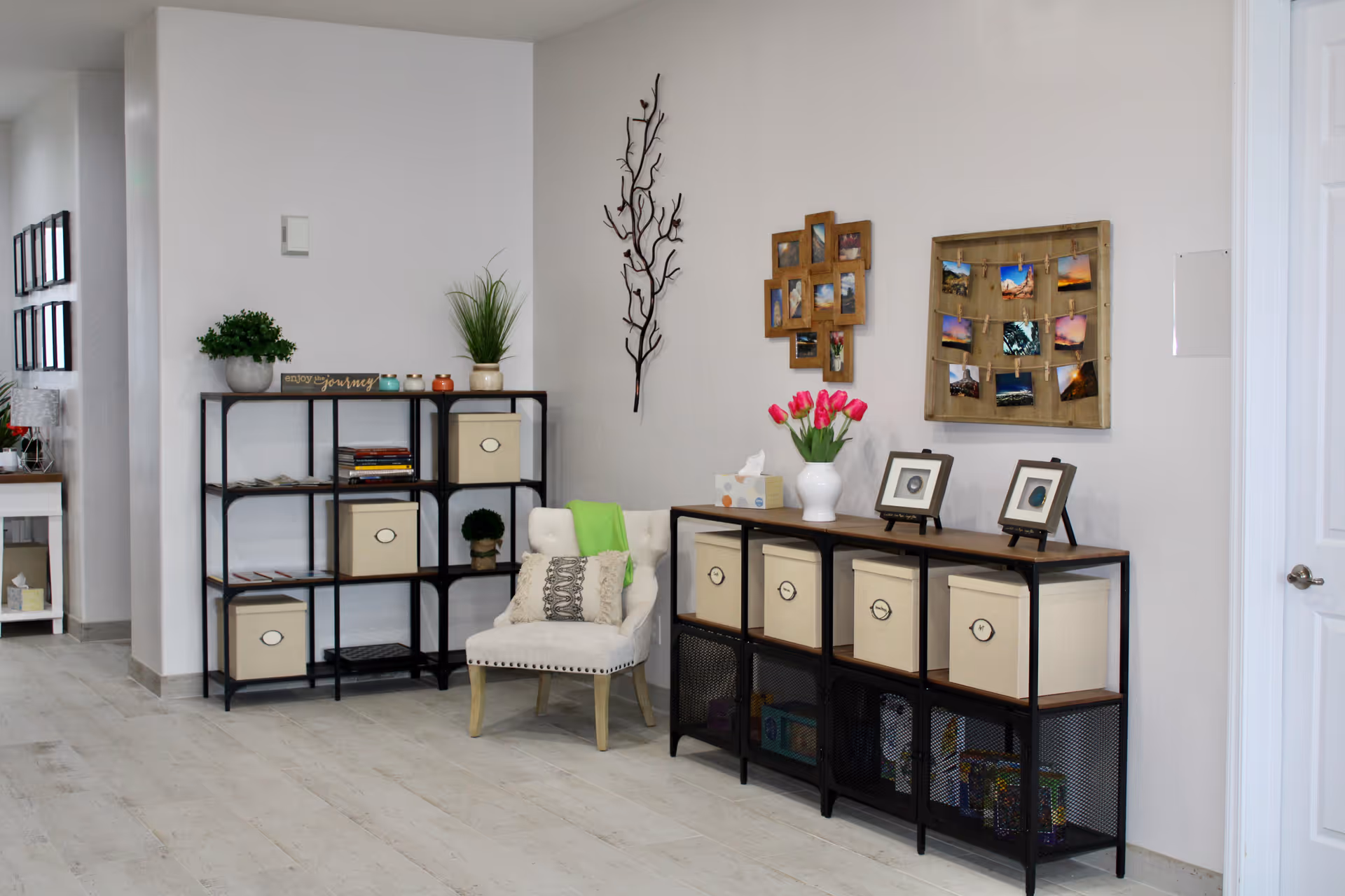 A cozy corner of an interior room featuring two black metal shelving units with wooden tops and beige storage boxes. One shelving unit has plants, books, and decorative items, while the other has framed photos, a tissue box, and a vase with pink tulips. A cushioned chair with a decorative pillow and a green throw is placed between the shelves. The walls are decorated with a metal branch sculpture and two photo displays. The floor is light-colored wood, and a white door is visible on the right.