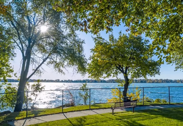 A peaceful outdoor scene at River Towers featuring a paved walkway along a waterfront with a metal fence. There are green trees providing shade, a bench near the fence, and the sun shining through the branches, reflecting off the water.