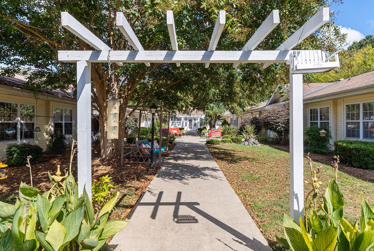A sunny outdoor courtyard at Langston Square featuring a white wooden pergola over a concrete pathway. The pathway is flanked by green plants, bushes, and trees, with benches and seating areas visible along the sides. Residential buildings with multiple windows surround the courtyard.