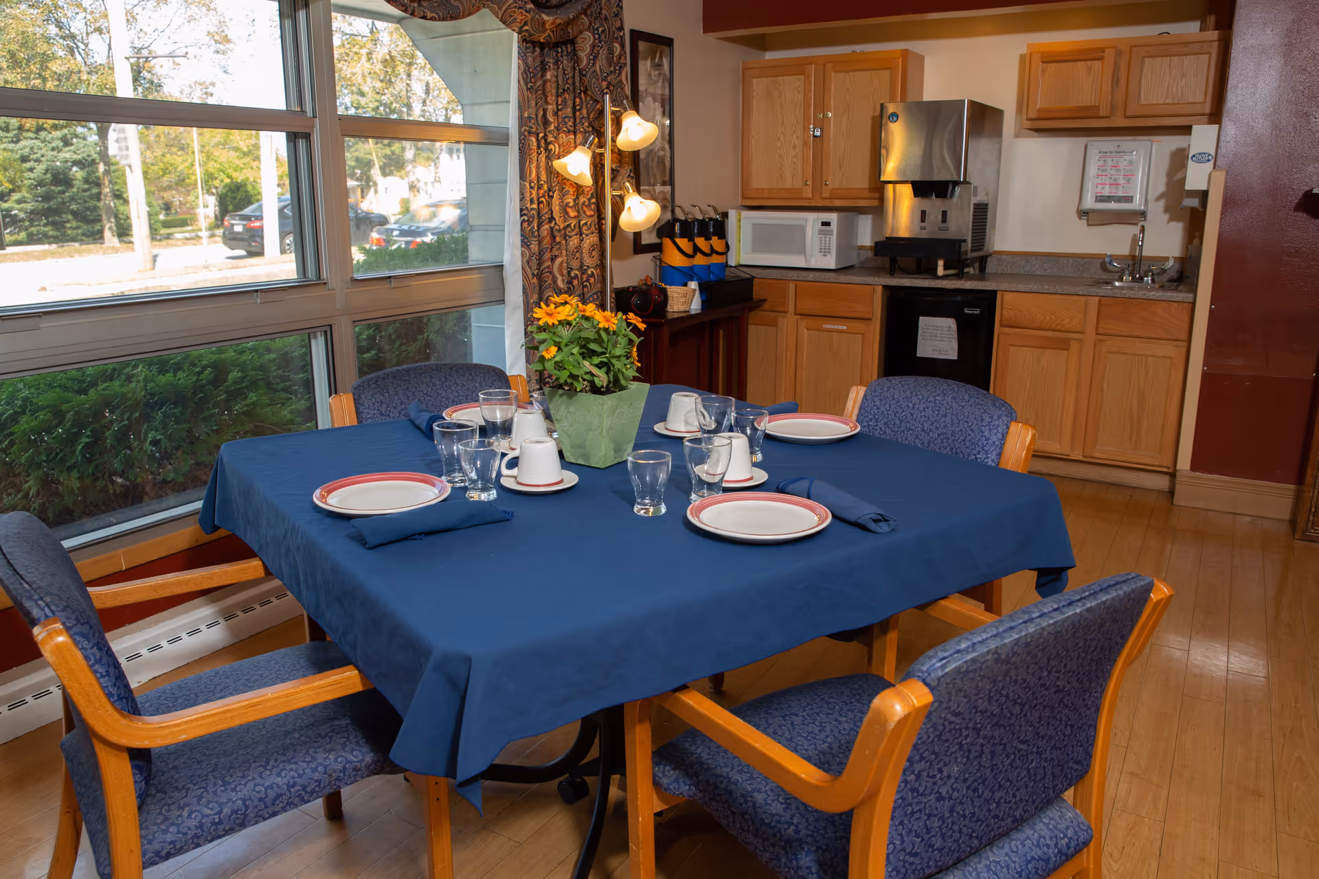 A dining area with a square table covered with a blue tablecloth set for four people with plates, cups, glasses, and napkins. The room has large windows with a view of greenery and parked cars outside. In the background, there is a kitchenette with wooden cabinets, a microwave, a coffee machine, and a small sink.