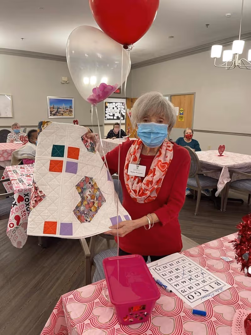 An elderly woman wearing a red shirt, heart-patterned scarf, and blue face mask is holding up a quilt with colorful patches in a decorated room. She is standing next to a table covered with a pink heart-themed tablecloth, a pink basket, and a bingo card. Other elderly people are seated at tables in the background, and there are balloons and Valentine-themed decorations around the room.