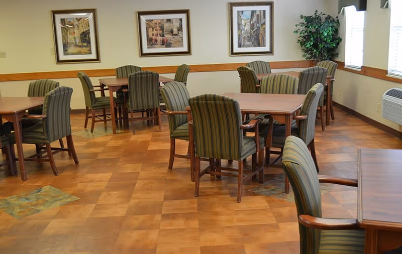 A dining room with several wooden tables and green striped upholstered chairs arranged neatly. The room has a wooden floor with a patterned design, three framed paintings on the wall, a potted plant in the corner, and windows with blinds allowing natural light to enter.