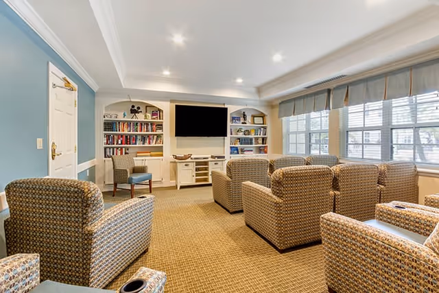 A cozy living room area with multiple patterned armchairs arranged facing a wall-mounted flat-screen TV. The room features built-in white bookshelves filled with books and decorative items on either side of the TV. Large windows with blinds allow natural light to fill the space, and the walls are painted in a soft blue and white color scheme.