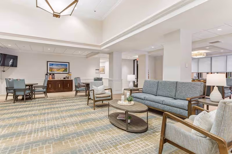 A spacious and well-lit common area in a senior living facility featuring a blue sofa, two armchairs with cushions, a round wooden coffee table with decorative items, and a side table with a lamp. In the background, there are dining tables with chairs, a wooden cabinet with coffee dispensers, a wall-mounted TV, and framed artwork. The room has a patterned carpet and white walls with recessed lighting.