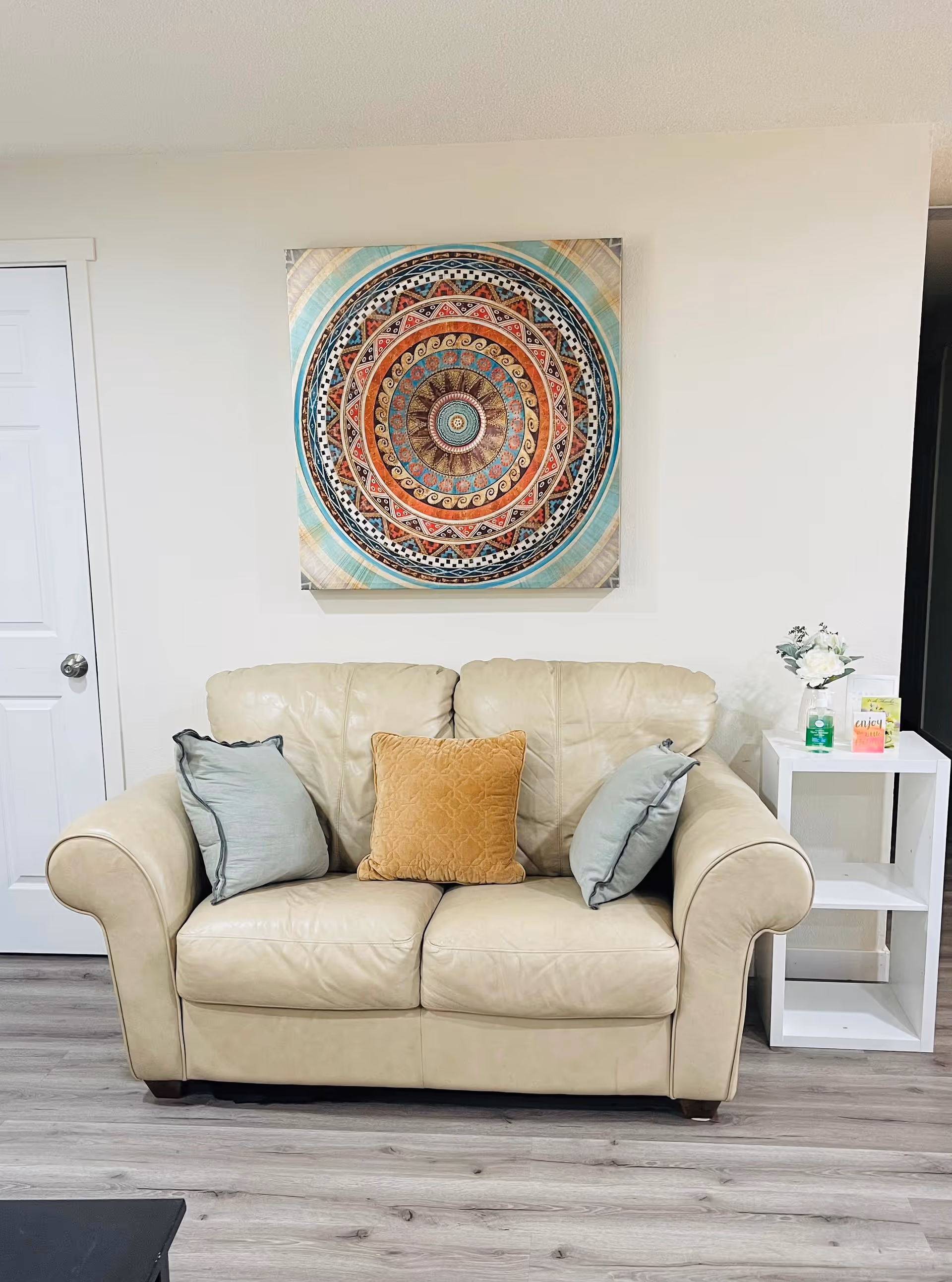 A beige leather loveseat with three decorative pillows, two light blue and one mustard yellow, placed against a white wall. Above the loveseat hangs a colorful circular mandala artwork. To the right of the loveseat is a white side table with a small flower arrangement, a bottle of hand sanitizer, and a card. The floor is light wood, and a white door with a silver knob is visible on the left side of the image.