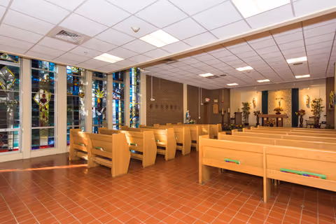 Interior view of a chapel or worship room with wooden pews arranged in rows facing an altar. The room has stained glass windows on one side and a tiled floor. The altar area is decorated with flowers and religious statues.