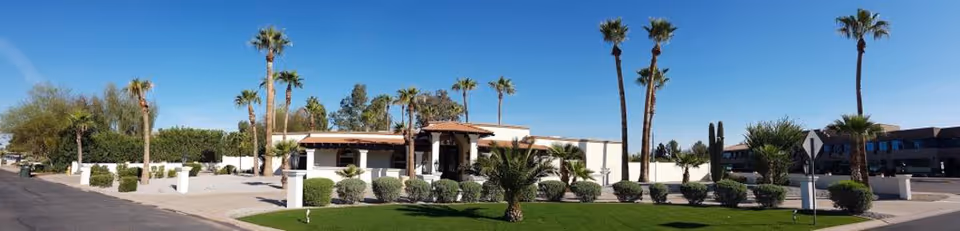 Wide panoramic view of a single-story building with a tiled roof surrounded by palm trees and well-maintained landscaping including bushes and green grass under a clear blue sky.