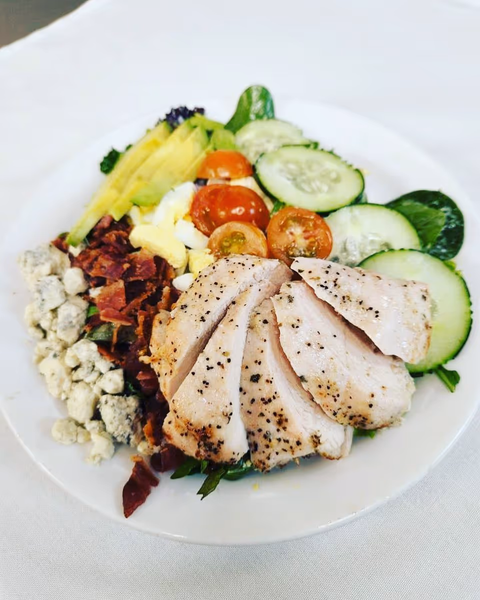 A plate of salad with sliced grilled chicken breast, cherry tomatoes, cucumber slices, avocado, bacon bits, blue cheese crumbles, and chopped boiled egg on a bed of mixed greens.