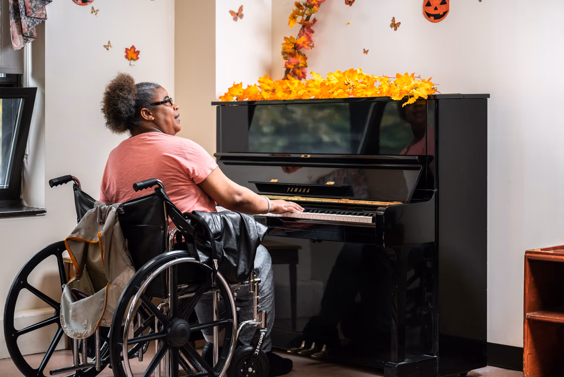A woman in a wheelchair playing a black upright piano decorated with autumn leaves and small butterfly and pumpkin wall decorations in a well-lit room.