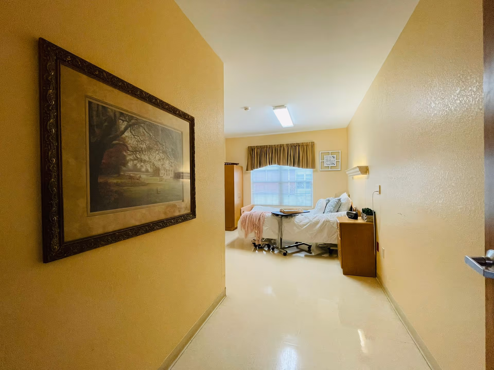 View down a hallway leading to a bedroom in a senior living facility. The bedroom has a hospital-style bed with white bedding and a pink throw blanket, a wooden nightstand with a small plant and clock, a window with a valance curtain, and a framed picture on the wall. The hallway wall features a large framed painting of a tree and house.
