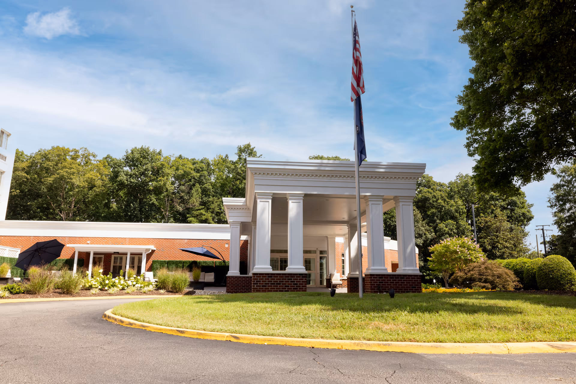 Front exterior view of The Barrington at Hioaks facility showing a covered entrance with white columns, an American flag on a flagpole, a circular driveway, and surrounding greenery including trees and bushes under a blue sky with some clouds.