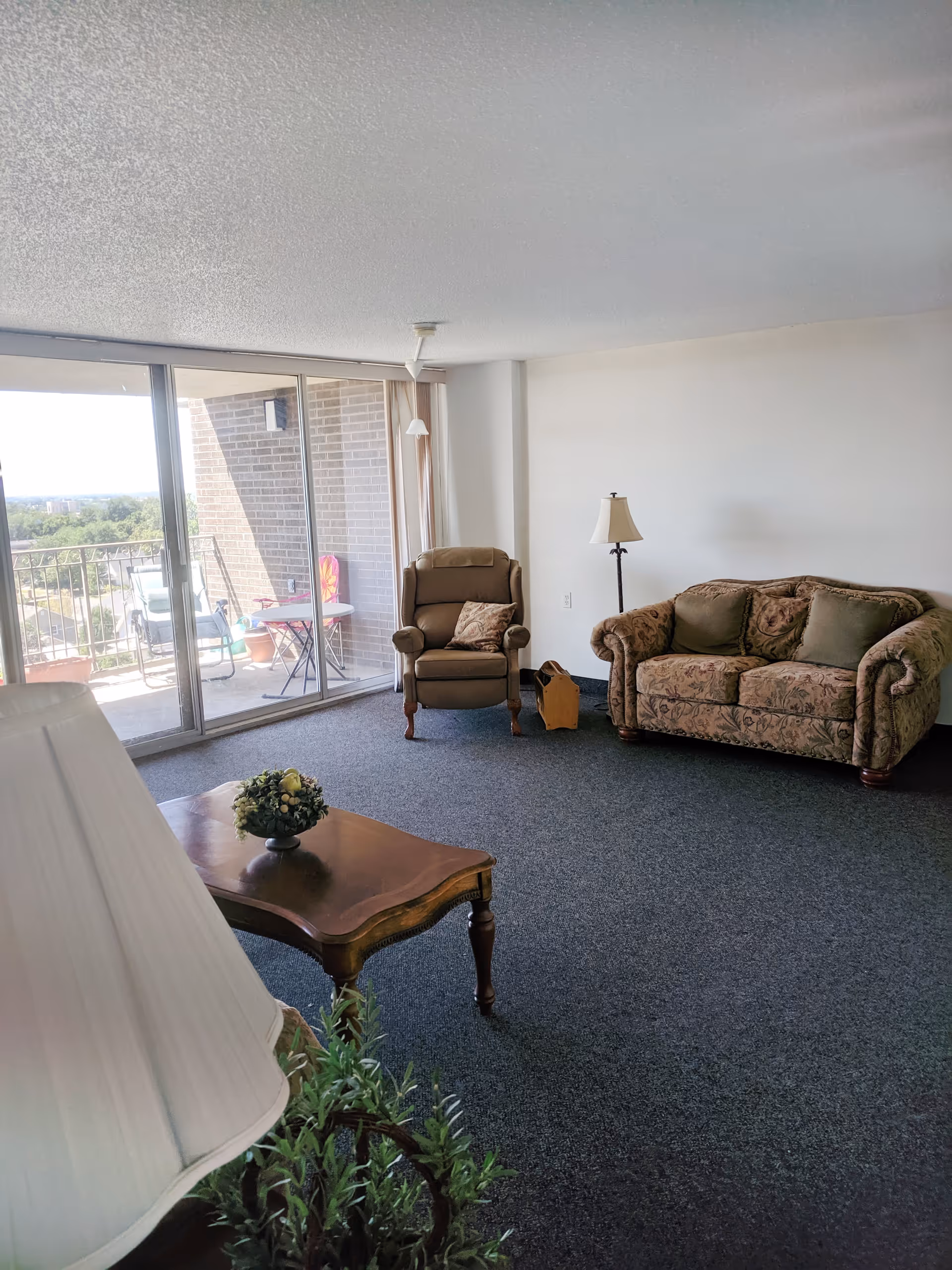 Bright living room with a patterned sofa and armchair, coffee table, floor lamp, and sliding glass doors opening to a balcony.