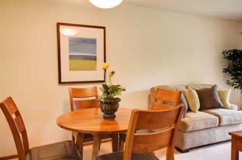 A cozy interior living space featuring a round wooden dining table with four wooden chairs. On the table is a small decorative plant. In the background, there is a beige sofa with multiple cushions and a framed abstract artwork hanging on the wall. A potted plant is visible in the corner of the room.