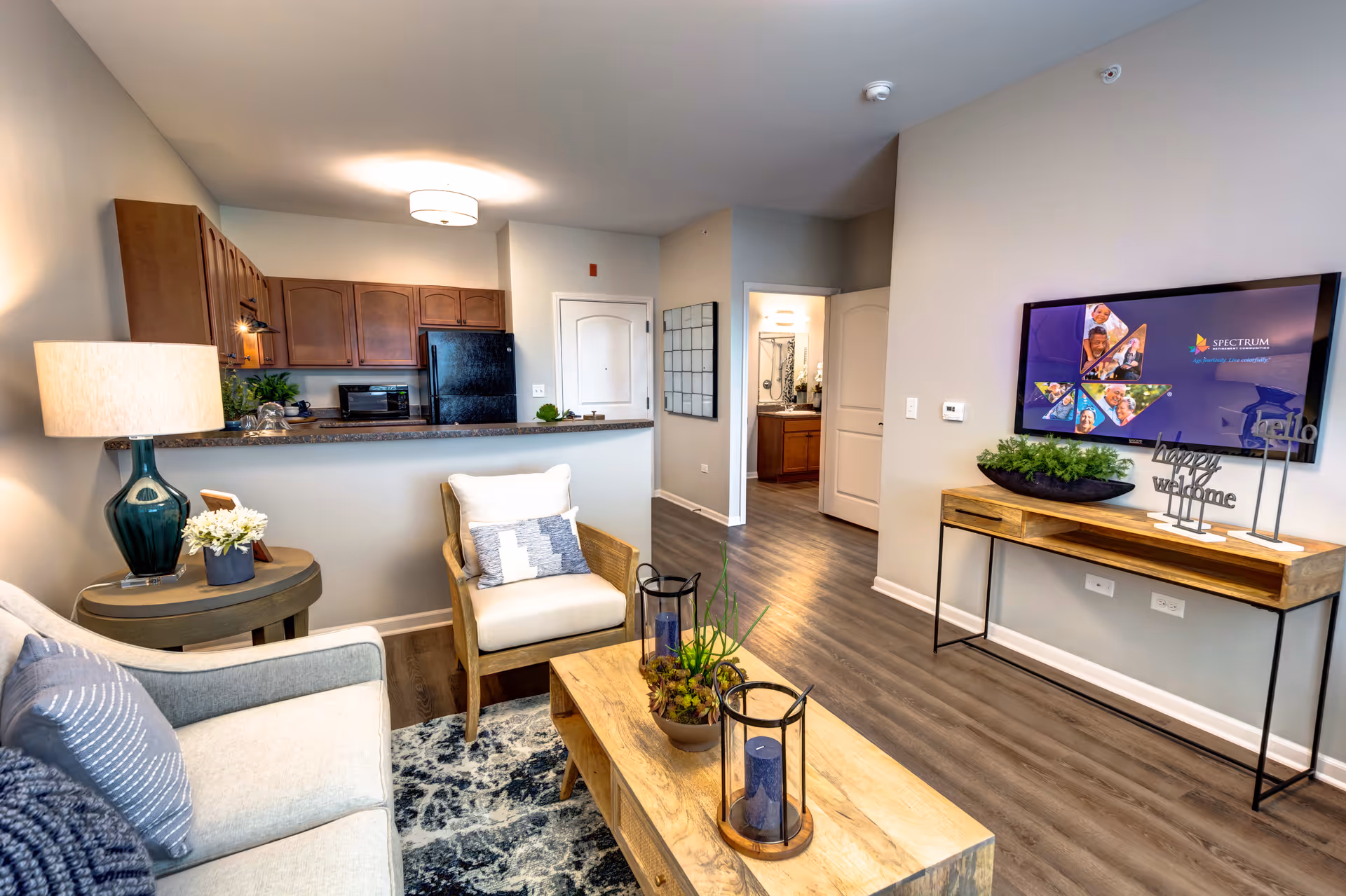 A cozy living room area in a senior living facility featuring a beige sofa with blue and gray pillows, a wooden armchair with a cushion, a wooden coffee table with decorative plants and candles, a side table with a lamp and flowers, and a wall-mounted TV displaying a welcome screen. The room has wood flooring and an open view into a kitchen with wooden cabinets and black appliances.