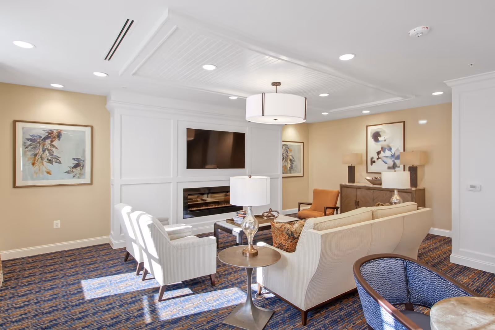 A bright and cozy senior living facility lounge with beige walls and blue patterned carpet. The room features a white sofa, two white armchairs, an orange armchair, a wooden sideboard with lamps, framed artwork on the walls, a mounted flat-screen TV above a modern electric fireplace, and a round ceiling light fixture.