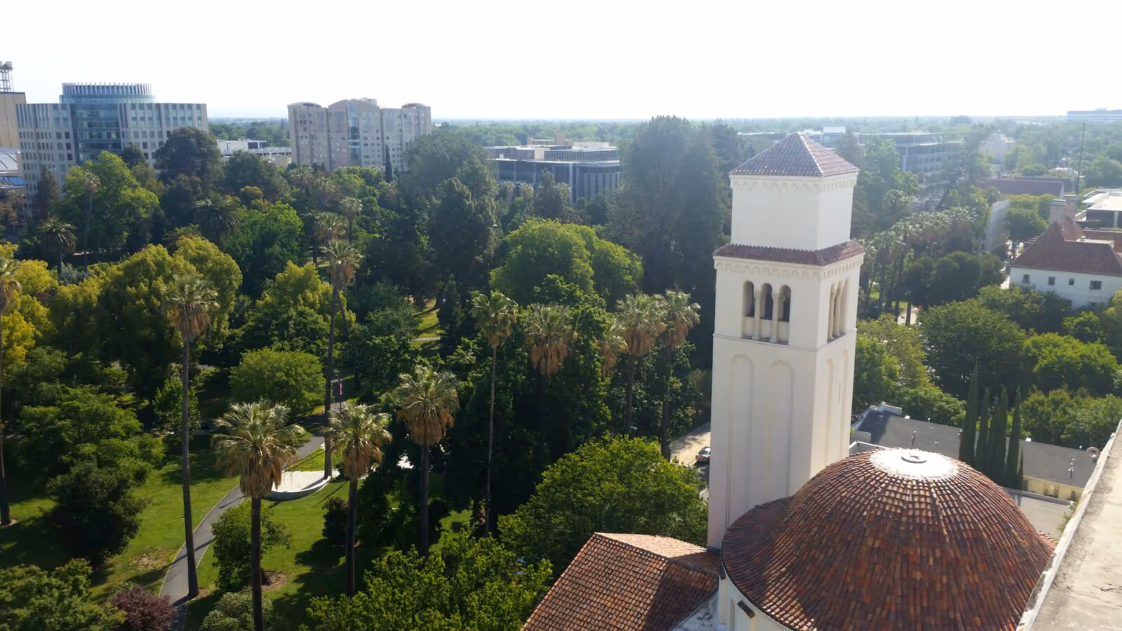 Aerial view of a green park area with tall palm trees and other lush trees, adjacent to a white building with a tall tower and a red-tiled dome roof. In the background, there are several modern buildings under a clear sky.