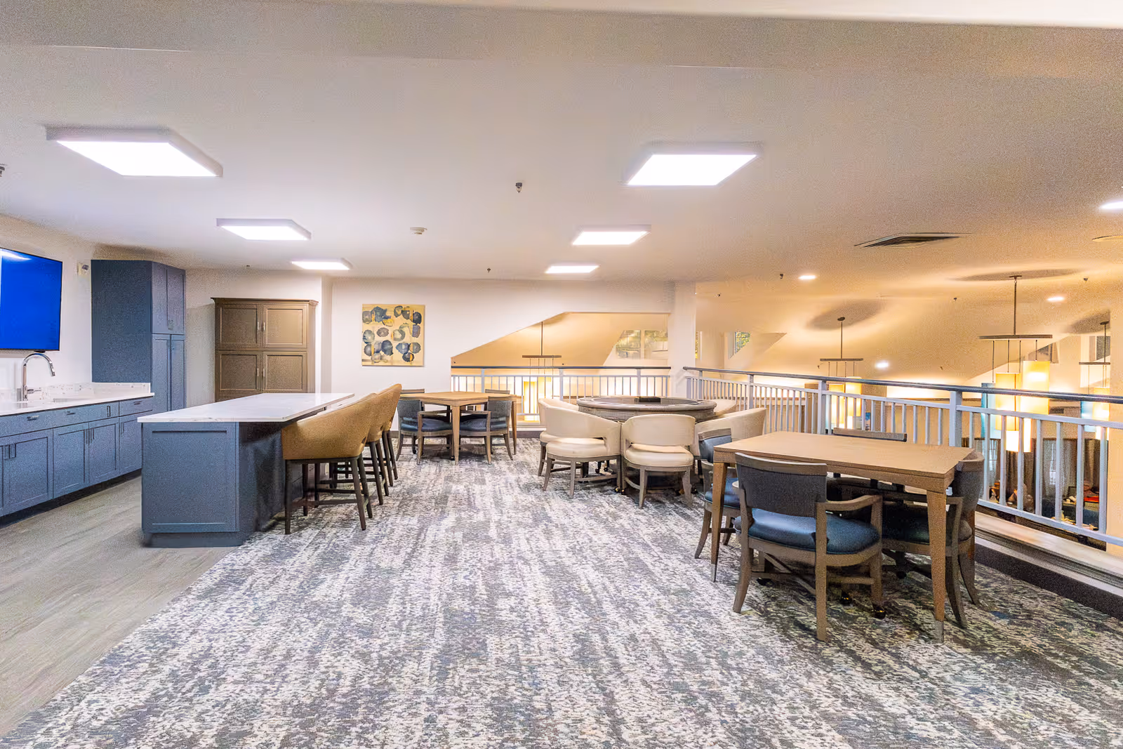 A spacious interior common area with a kitchen island featuring bar stools on the left, multiple tables with chairs arranged for seating, and modern lighting fixtures overhead. The area has a carpeted floor and overlooks a lower level through a railing.