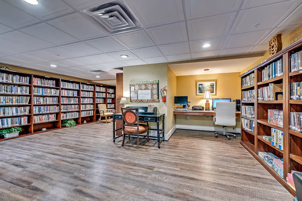 Interior view of a library or reading room in a senior living facility with wooden bookshelves filled with books and board games. There are two desks with chairs, one with a lamp and bulletin board, and the other with two computer monitors and a lamp. The floor is wooden, and the ceiling has recessed lighting.