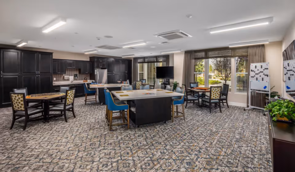 A spacious senior living common area with multiple tables and chairs arranged for games and socializing. The room features a large kitchen area with dark cabinetry and stainless steel appliances. There are large windows with curtains allowing natural light to brighten the space, and a television mounted on a stand near the windows. The carpet has a patterned design, and there are plants and whiteboards with crossword puzzles on the right side of the room.