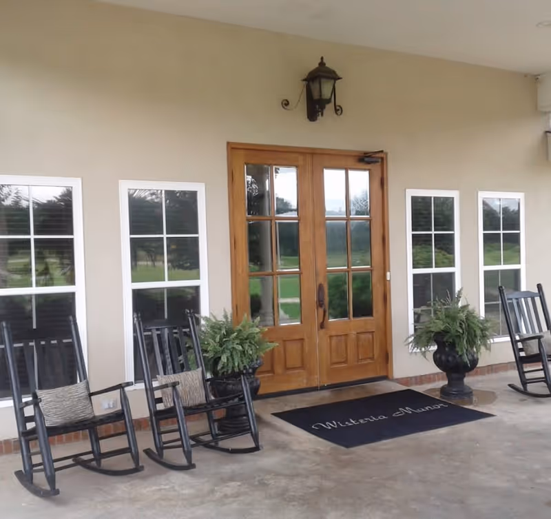 Entrance of Wisteria Manor Personal Care Home featuring double wooden doors with glass panels, flanked by four windows. There are black rocking chairs with cushions and two large potted plants on either side of the door. A black mat with 'Wisteria Manor' written on it lies in front of the doors, and a wall-mounted lantern light fixture is above the doors.
