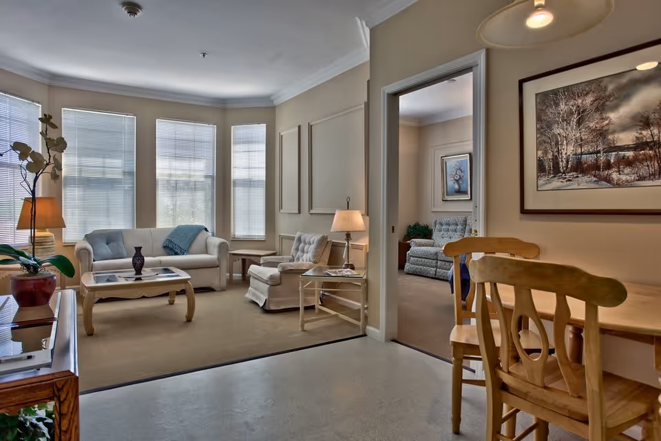 A cozy living room area with a beige sofa, an armchair, a coffee table with a decorative vase, and a side table with a lamp. Large windows with blinds let in natural light. Adjacent to the living room is a small dining area with a wooden table and chairs. A doorway leads to another room with additional seating and framed artwork on the walls.
