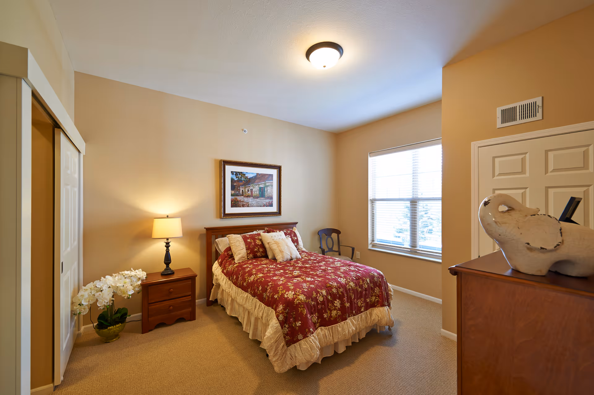 A cozy bedroom with beige walls and carpet, featuring a bed with a red floral bedspread and multiple pillows. There is a wooden nightstand with a lamp and a potted white orchid next to it. A framed painting hangs above the bed. A wooden chair is placed near a window with blinds, and a wooden dresser with a decorative elephant sculpture is visible in the foreground.