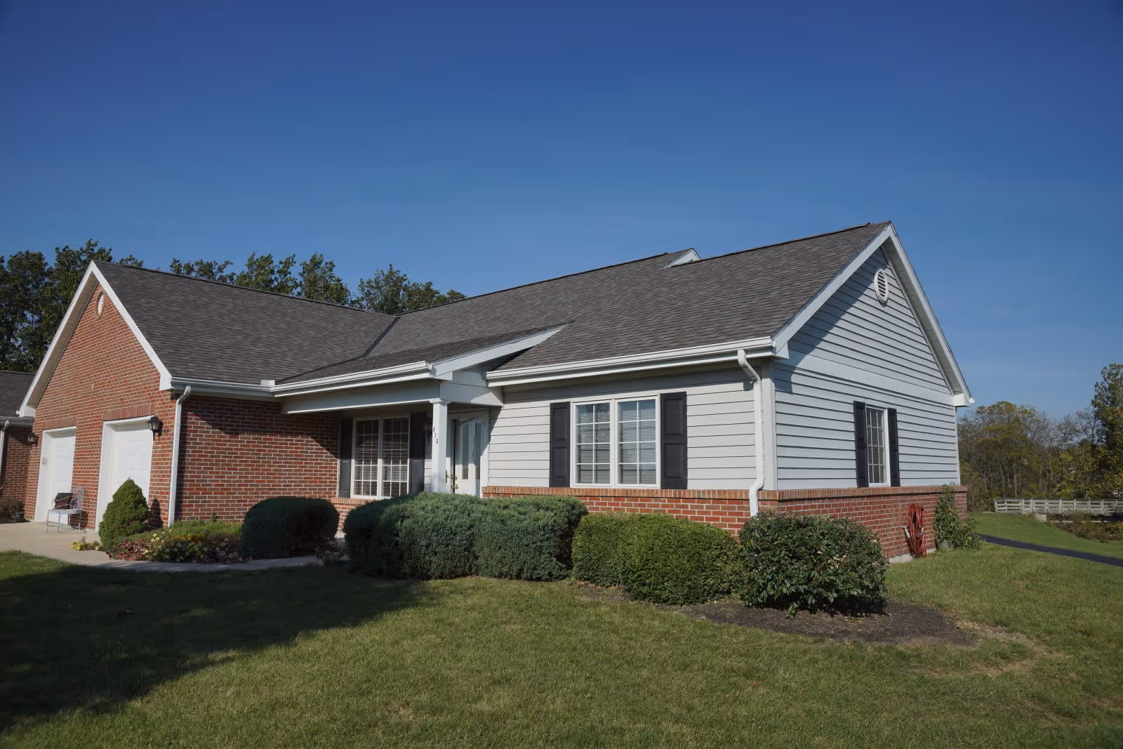 Exterior view of a single-story residential building with a combination of red brick and light gray siding, black shutters on the windows, a dark shingled roof, and a well-maintained lawn with shrubs in front under a clear blue sky.