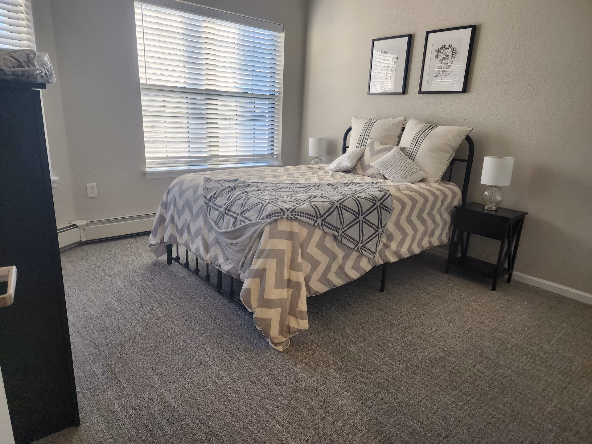 A bedroom with a bed covered in a chevron patterned blanket and multiple pillows. There are two framed pictures on the wall above the bed, a window with blinds letting in natural light, a nightstand with a lamp on the right side of the bed, and a dresser partially visible on the left side.