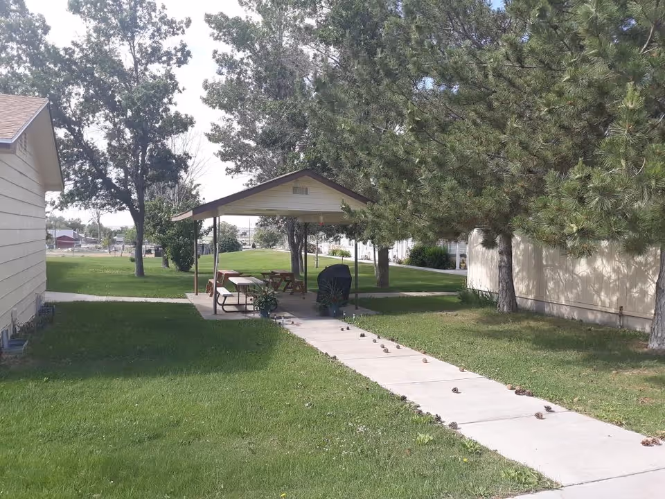 Outdoor area at GoodLife Senior Living & Memory Care of Elmo, UT featuring a covered picnic area with benches and a grill, surrounded by green grass, trees, and buildings.