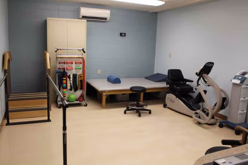 A small physical therapy room with a set of wooden stairs with handrails, a therapy table with cushions, a black rolling stool, a recumbent exercise bike, and a rack holding various therapy bands and equipment. The walls are painted light blue and white, and there is an air conditioning unit mounted on the wall.