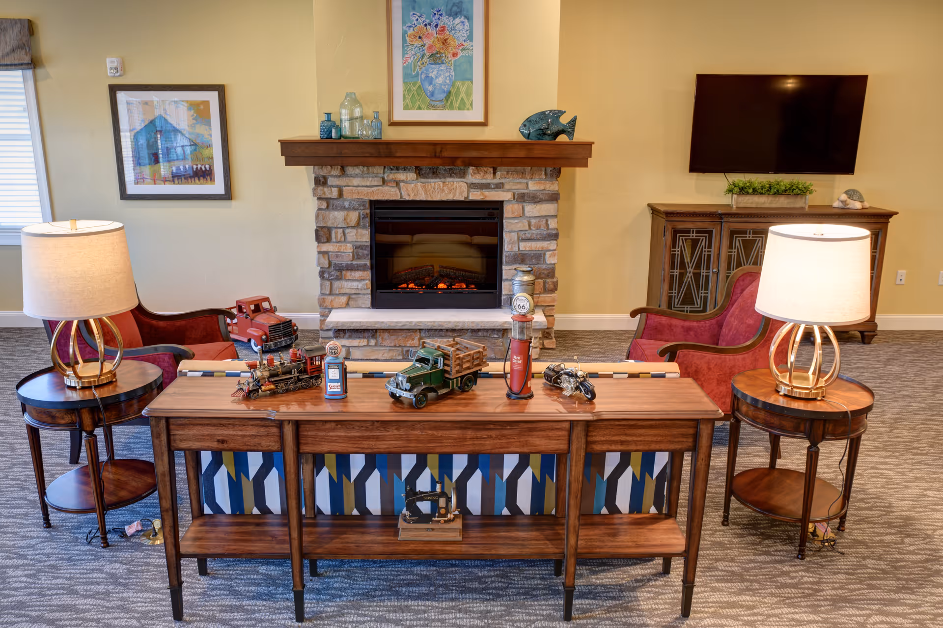 A cozy living room area with a stone fireplace in the center, flanked by two red armchairs. A wooden console table in front of the fireplace displays vintage toy vehicles and decorative items. Two round side tables with lamps are positioned beside the armchairs. A flat-screen TV is mounted on the wall above a wooden cabinet. The walls are painted light yellow, and there is framed artwork above the fireplace and on the left wall.