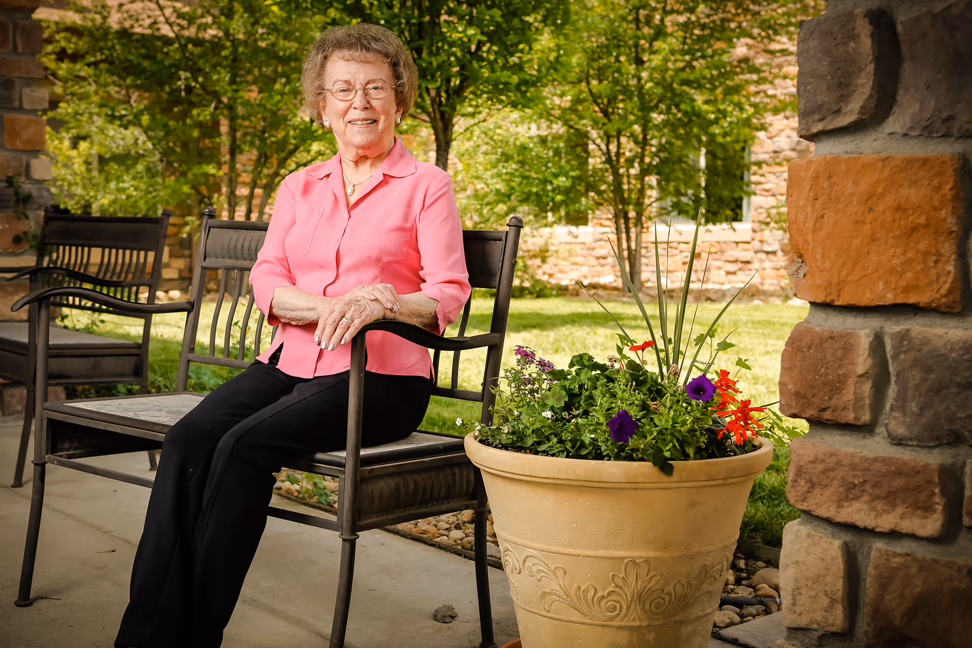 An elderly woman wearing a pink blouse and black pants sits on a metal bench outdoors. She is smiling and has short curly hair and glasses. Next to her is a large decorative flower pot with colorful flowers. The background shows green trees and a stone building wall.