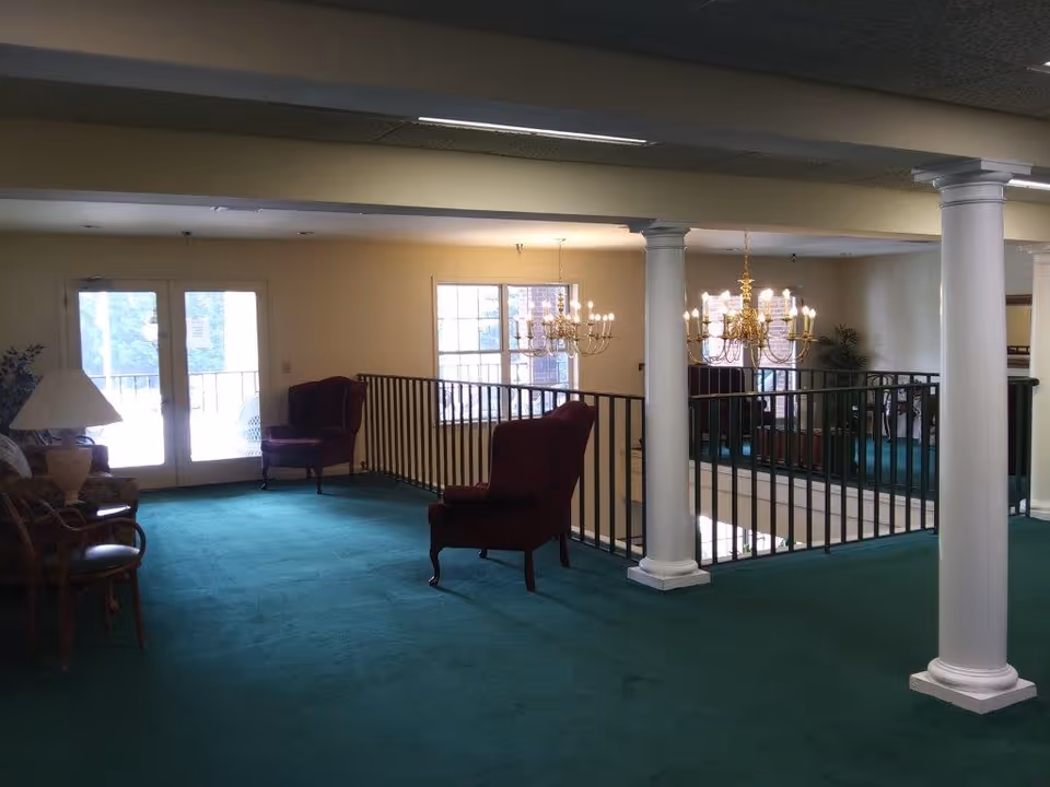 Upper-level lounge with green carpet, wingback chairs, white columns and a railing overlooking a chandelier-lit lower level and glass doors to a balcony.