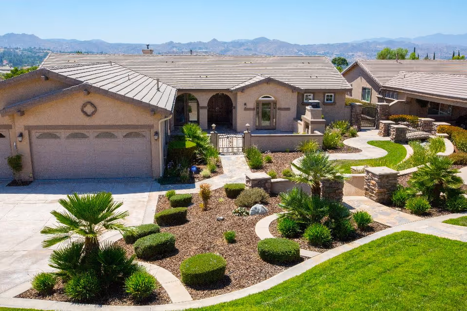 Front exterior view of a single-story senior living facility building with a tiled roof, garage, landscaped garden with trimmed bushes and palm trees, paved walkways, and a gated entrance. Mountains are visible in the background under a clear blue sky.