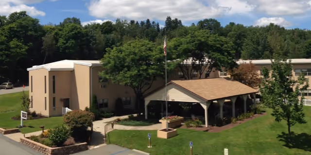 Exterior view of Forest City Nursing And Rehab Center showing a two-story beige building surrounded by green trees and a well-maintained lawn. There is a covered pavilion with a flagpole in front and a parking area with handicap parking spaces visible.