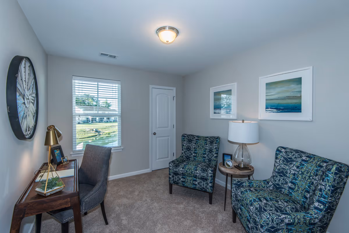 A small, cozy room with beige walls and carpeted floor. The room features a wooden desk with a gray upholstered chair, a gold desk lamp, and decorative items including a small plant and framed pictures. On the opposite side, there are two patterned armchairs with a small round wooden side table between them, holding a glass lamp and a clock. Two framed abstract paintings hang on the wall above the chairs. A window with white blinds lets in natural light, and a white door is visible in the corner.