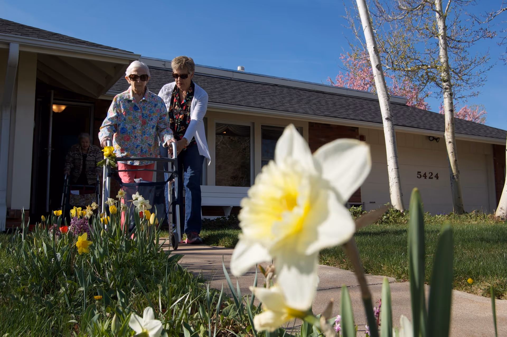 An elderly woman using a walker is being assisted by a caregiver as they walk on a sidewalk outside a single-story residential building with the number 5424. There are blooming flowers and trees in the foreground and background under a clear blue sky.