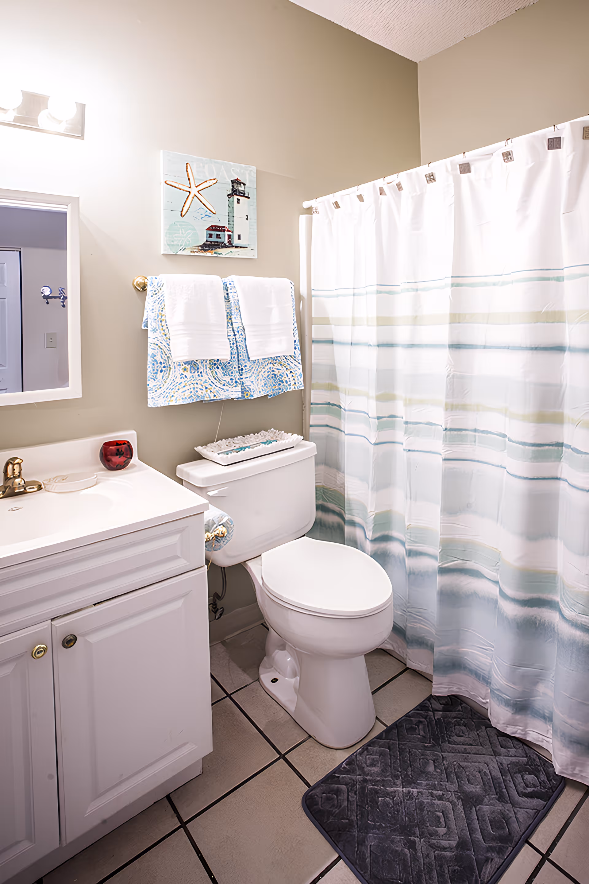 A clean bathroom featuring a white toilet, a white vanity with a sink and gold faucet, a mirror above the sink, and a shower with a white curtain that has blue and green stripes. Two white towels hang on a towel rack above the toilet, and a decorative picture with a starfish and lighthouse is mounted on the wall. A dark gray bath mat is placed on the tiled floor.