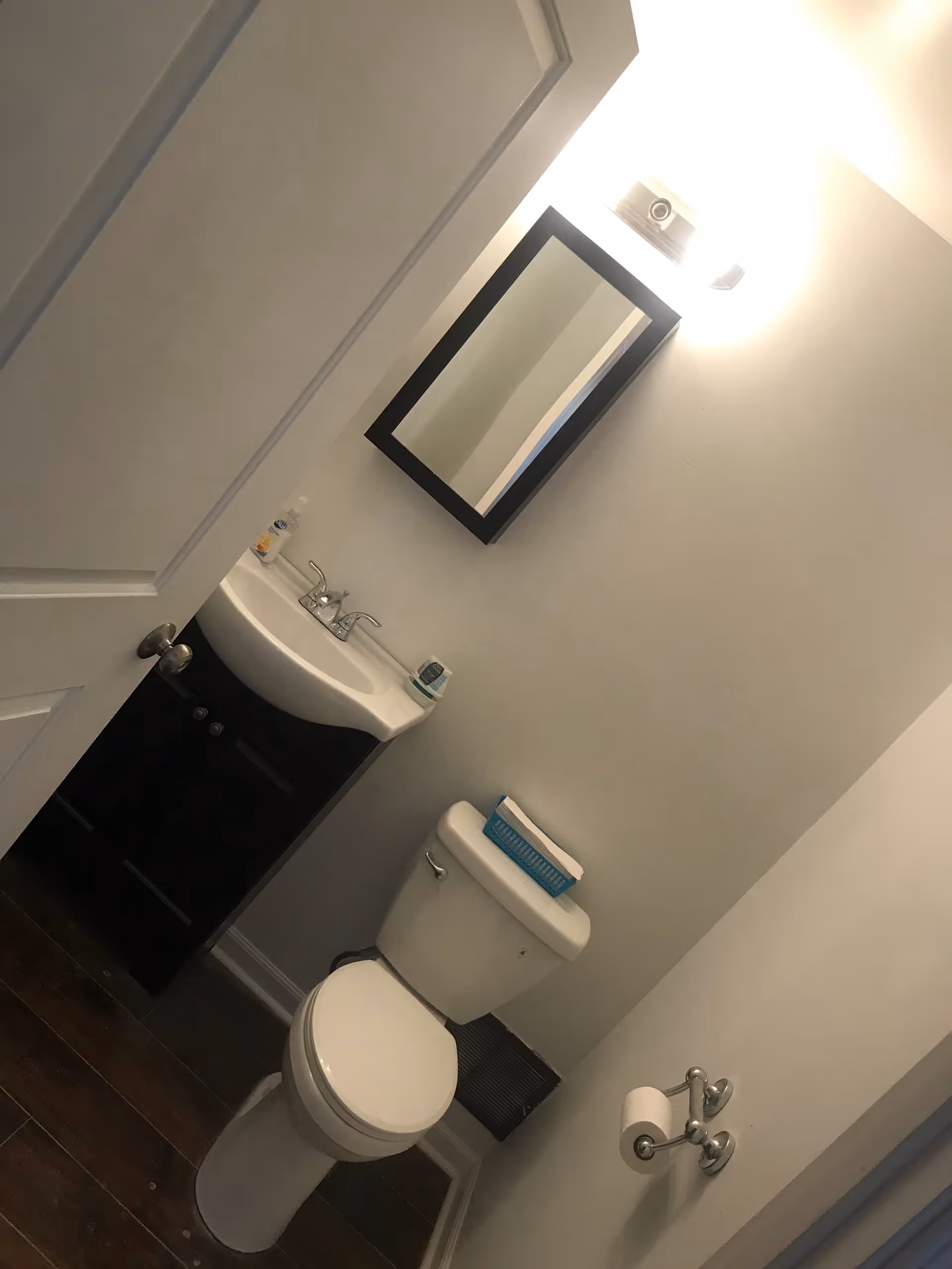 Small bathroom with a white toilet and a black vanity with a white sink. Above the sink is a rectangular mirror with a black frame and a light fixture above it. There is a toilet paper holder mounted on the wall and a small blue basket on top of the toilet tank.