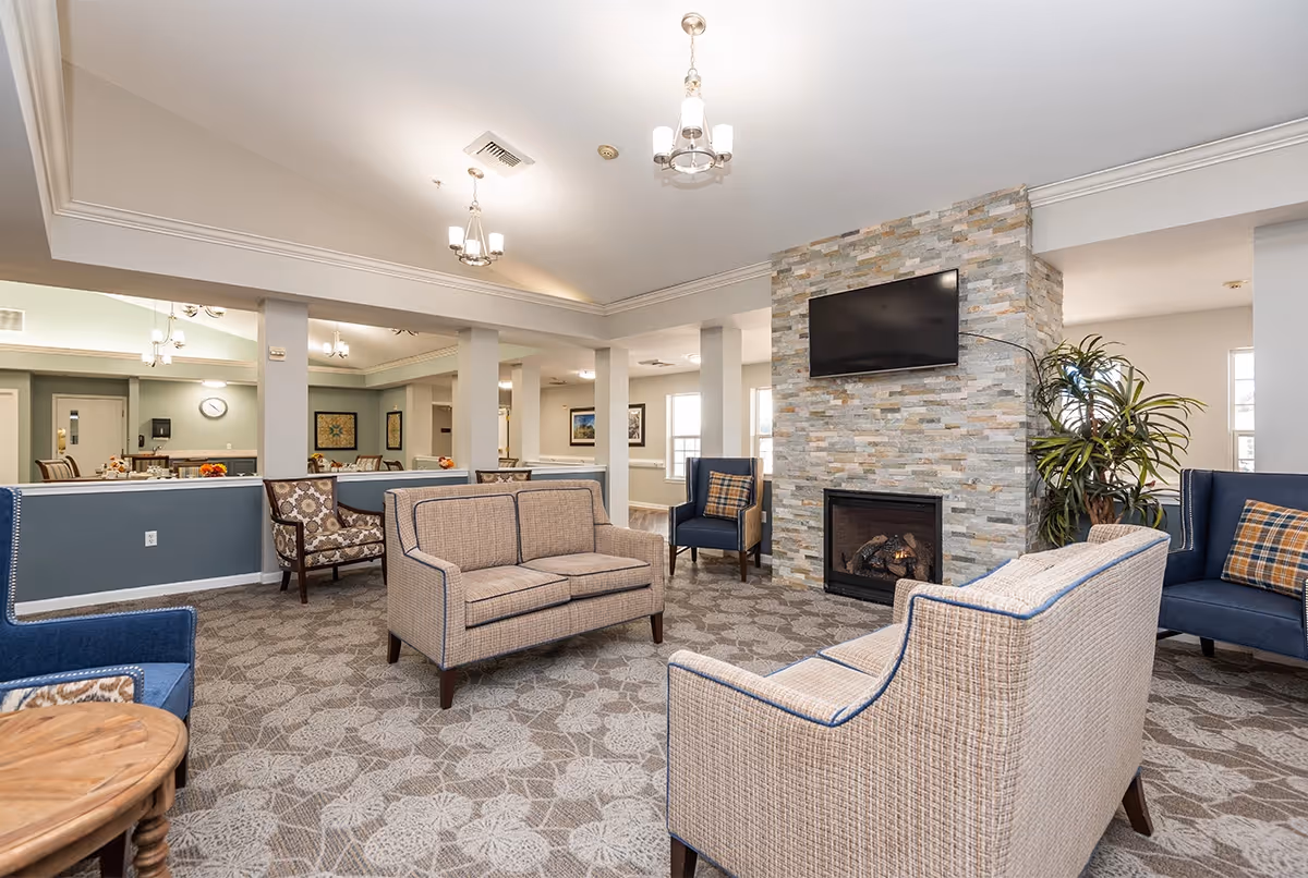 A spacious senior living facility common area with beige and blue upholstered sofas and armchairs arranged around a stone fireplace with a mounted flat-screen TV. The room has patterned carpet, light-colored walls, and multiple ceiling light fixtures. In the background, there is a dining area with tables and chairs.